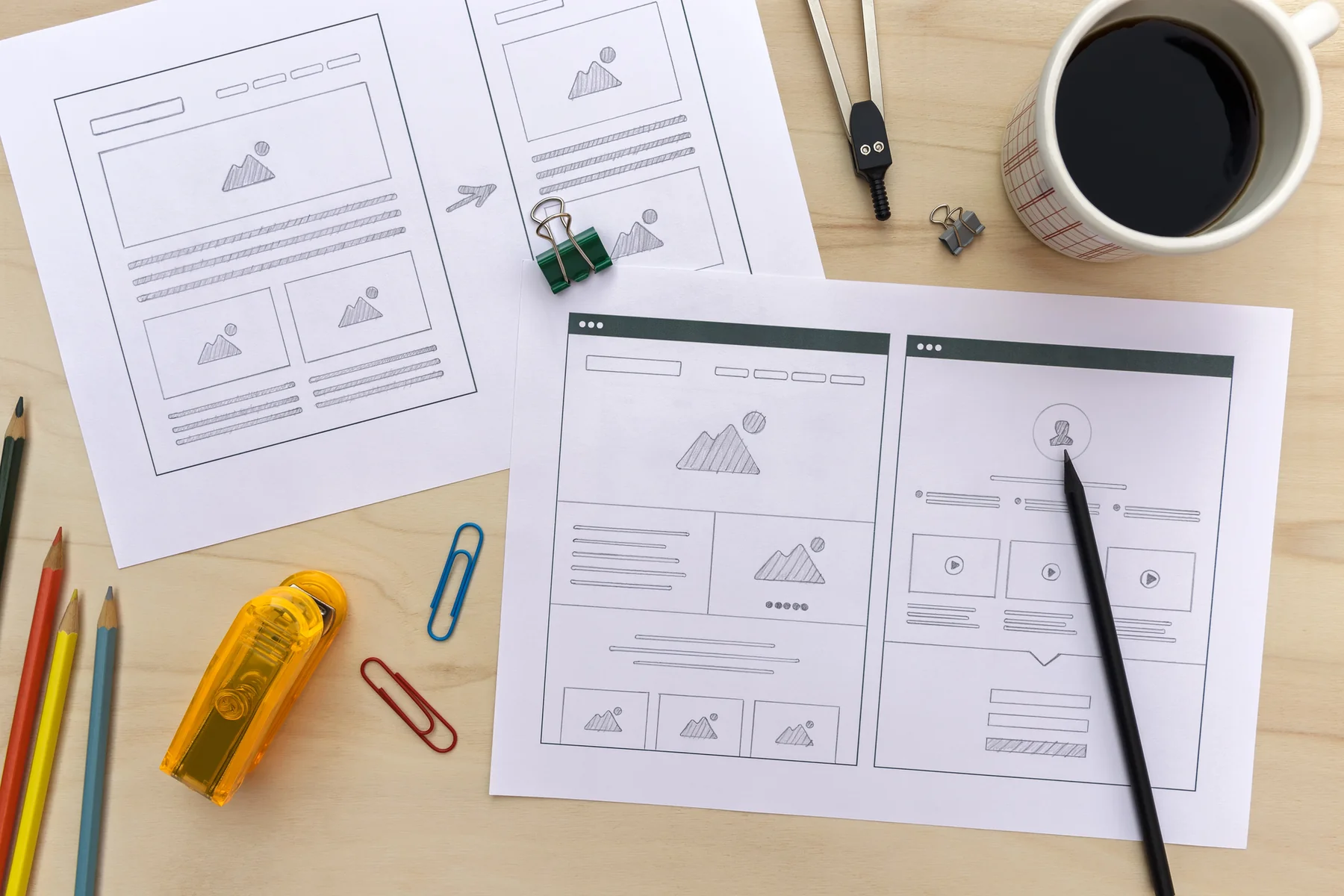 Paper website layout sketches, pencils and a cup of coffee on a desk.